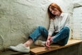 Woman with red hair in jeans, sneakers and white shirt sits on wooden bench against background of roughly plastered wall