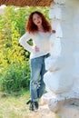 A woman model with red hair and a white knitted sweater stands behind the wall of an old house with a thatched brown roof Royalty Free Stock Photo