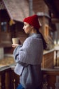 woman in a red hat and scarf and a mug stands at a wooden house in the woods Royalty Free Stock Photo