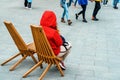 A woman in a red jacket sits on a wooden chair in a public place. Rest in the park during the cool season. Unrecognizable person.