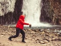 A woman in a red jacket stands against the backdrop of a stormy waterfall flowing down from wet icy rocks filling her