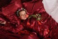 Woman in red pajamas lying on a bed on silk red linen with hair curlers and a rose in her hand Royalty Free Stock Photo