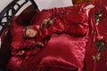 Woman in red pajamas lying on a bed on silk red linen with hair curlers and a rose in her hand Royalty Free Stock Photo