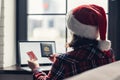 Woman in a red santa claus hat holding credit card and american passport using laptop for making order sitting near window. Royalty Free Stock Photo