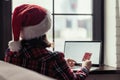 Woman in a red santa claus hat holding credit card using laptop for making order sitting near window. Christmas online shopping. Royalty Free Stock Photo