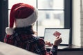 Woman in a red santa claus hat holding credit card using laptop for making order sitting near window. Christmas online shopping. Royalty Free Stock Photo