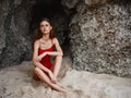 Woman in red swimsuit sitting by the rocks on the sand with a beautiful tan from the sun on the beach, travel to Bali, a Royalty Free Stock Photo