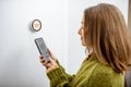 Woman regulating heating temperature with phone and thermostat at home Royalty Free Stock Photo