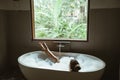 Woman relaxing in foam bath with bubbles in dark bathroom by window