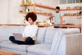 Woman Relaxing Sitting On Sofa At Home Using Laptop Computer With Man In Kitchen Pouring Drink