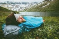 Woman relaxing in sleeping bag laying on grass enjoying lake and mountains