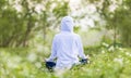 Rear view of woman in hoodie is relaxingly practicing meditation yoga in forest full of daisy flower in summer to attain happiness