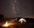 Woman resting at night camping near campfire, tourist tent, bicycle under evening sky full of stars Royalty Free Stock Photo