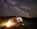 Woman resting at night camping near campfire, tourist tent, bicycle under evening sky full of stars Royalty Free Stock Photo