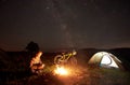 Woman resting at night camping near campfire, tourist tent, bicycle under evening sky full of stars