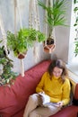 Woman resting sitting on couch under cotton macrame plant hanger with houseplants reading magazine