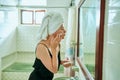 Woman in retro bathroom applies face cream, wrapped in towel. Skin care routine, mirror reflection at home spa. Female