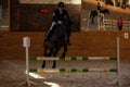 Woman rider on a black steed overcomes the barrier indoors.