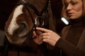 Woman rider putting bridle horsey muzzle while standing in stable