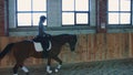 View of woman training and riding horse on sandy arena under roof.