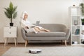 Woman running robot vacuum while resting with laptop on sofa