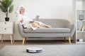 Woman running robot vacuum while resting with laptop on sofa