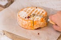 Woman`s hand cutting whole wheel of soft French, German cheese with orange rind with mold on parchment paper, wood cutting board