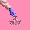 A woman`s hand in a disposable bright glove holds a shopping metal basket from a supermarket on a pink background. Royalty Free Stock Photo