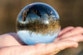 Woman`s hand holding Clear Quartz Sphere reflecting lake, forest, sky.