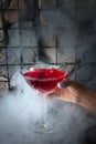 Woman`s hand holding a glass of red cocktail with a cherry. Around a lot of smoke from a hookah. Stone wall as background Royalty Free Stock Photo