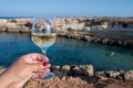 Woman`s hand holding glass of white dry white wine with view on rocks and blue sea bay water near Protaras touristic town on Royalty Free Stock Photo