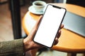 A woman`s hand holding mobile phone with blank white desktop screen with laptop and coffee cup on table Royalty Free Stock Photo