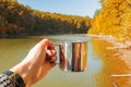 Woman`s hand holding a mug with hot tea near the lake in a warm sunny autumn day. Royalty Free Stock Photo