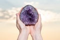 Woman`s hand holding partially polished Heart shaped Amethyst geode specimen from Brazil at sunrise in front of the lake