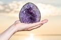 Woman`s hand holding partially polished Heart shaped Amethyst geode specimen from Brazil at sunrise in front of the lake