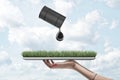 Woman`s hand holding patch of green lawn on metal tray with black barrel of oil tipped in the air spilling big drop of Royalty Free Stock Photo