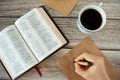 Woman`s hand holding pen to write on old vintage paper with an open Holy Bible Book, coffee cup, and envelope on a wooden retro ta