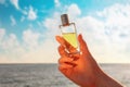 A woman& x27;s hand holds a glass perfume bottle against a blue sky with clouds and the ocean. The concept of perfumery