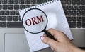 A woman`s hand holds a magnifying glass over the acronym ORM against the background of a laptop