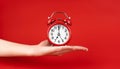 A woman\'s hand holds a red alarm clock against a red background