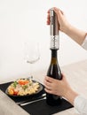 A woman`s hand opens a bottle of wine with an electric corkscrew made of gray metal. Next to it is a plate of pasta, a glass and Royalty Free Stock Photo