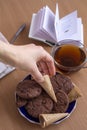 A woman`s hand takes a waffle tube from a saucer with sweets. There is a cup of tea and an open book on the table. Royalty Free Stock Photo