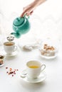 Woman`s hande pouring tea from teal colored teapot, white tea cups, sugar bowl standing on white table