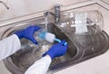 WomanÃ¢â¬â¢s hands with gloves washing dish with fresh water at kitchen faucet over the metal sink. Human washes glass jar. Close up