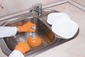 WomanÃ¢â¬â¢s hands with orange gloves cleaning sewer and water at kitchen faucet over metal sink. Female hand with plunger. Home