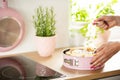 Woman`s hands sifting sugar on a sponge cake in a pastel pink springform pan in a bright kitchen interior