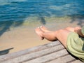 Woman's legs on the beach. Woman in casual green dress sit on wooden seat on sandy beach and blue sea background. Royalty Free Stock Photo