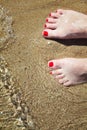 Woman`s pedicured feet with red nail polish on toes in the sand in water.