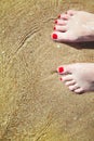 Woman`s pedicured feet with red nail polish on toes in the sand in water.