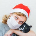 A Woman In A Santa Claus Hat And A Medical Mask Holds A Cute Black Cat In Her Hands.